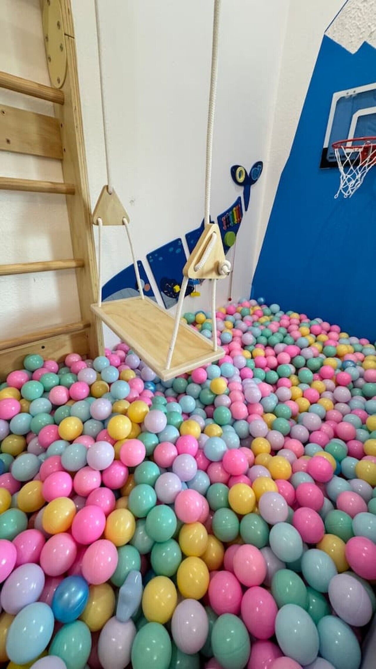 Una habitación con una piscina de pelotas multicolores, un columpio de madera y un aro de baloncesto.