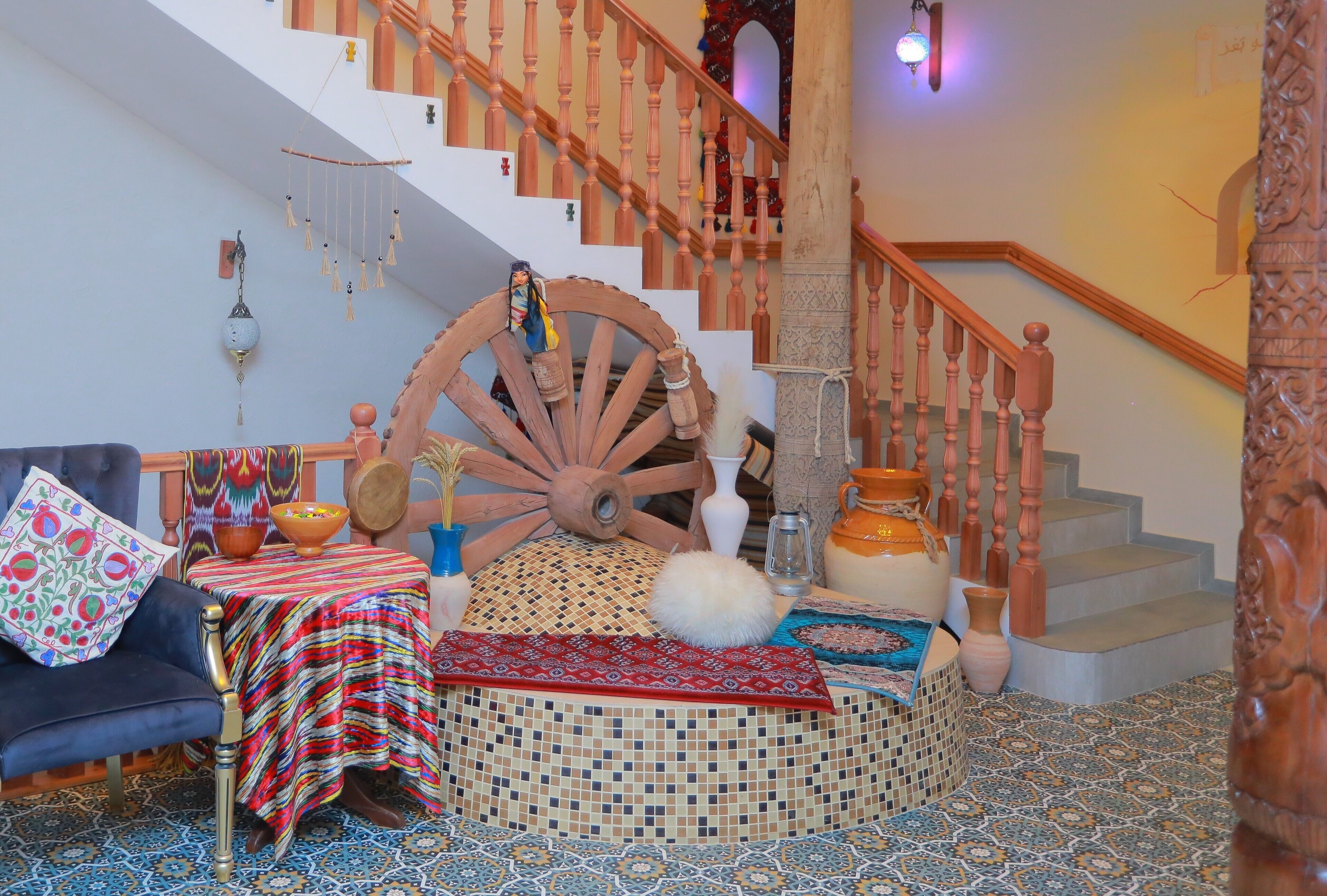 A room with a large wooden wheel, a staircase with wooden balusters, a patterned rug, and a blue-tiled floor.