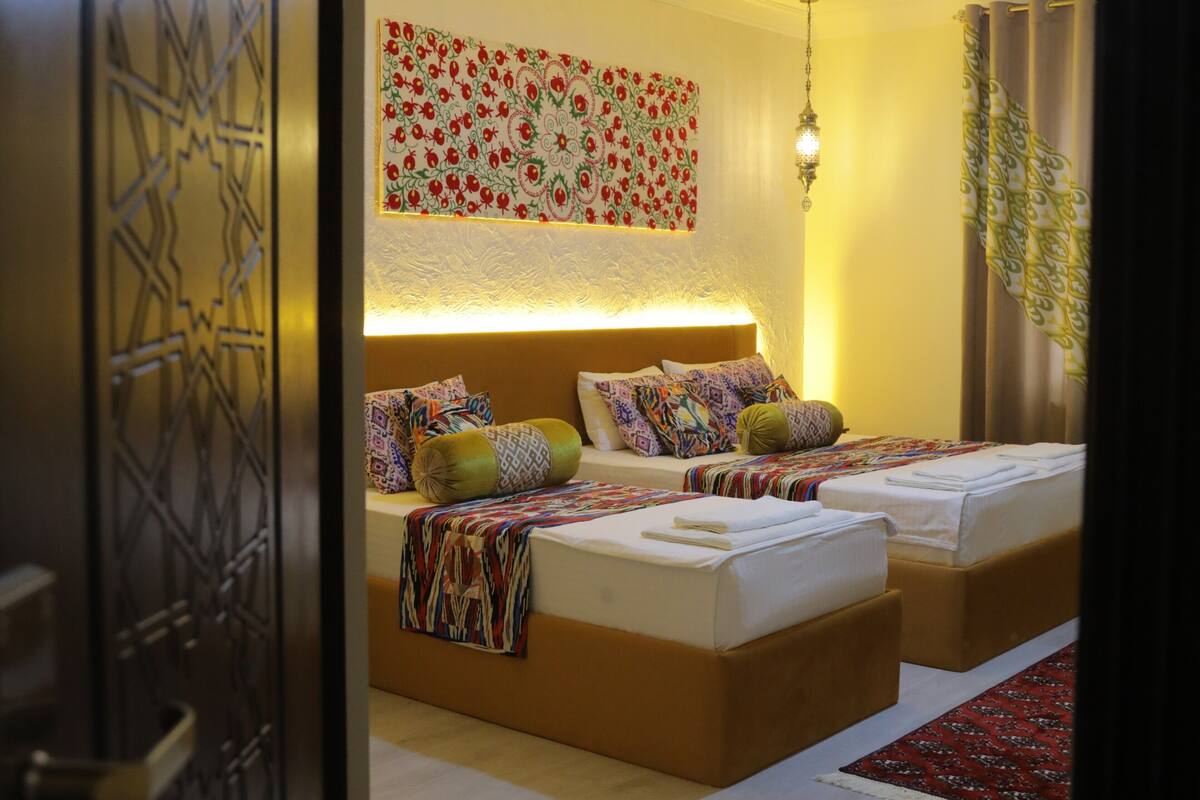 A hotel room with two beds, a patterned wall hanging, a hanging light fixture, and a patterned curtain.