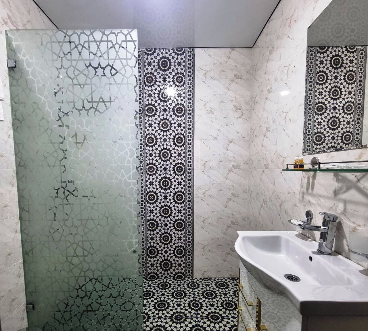 A bathroom with a glass shower enclosure, a sink with a mirror, and patterned tiles on the walls and floor.