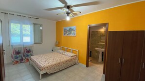 Een slaapkamer met een bed, een plafondventilator, een raam met zonwering, een radiator, een nachtkastje met een lamp, een aan de muur hangend schilderij en een houten garderobe.