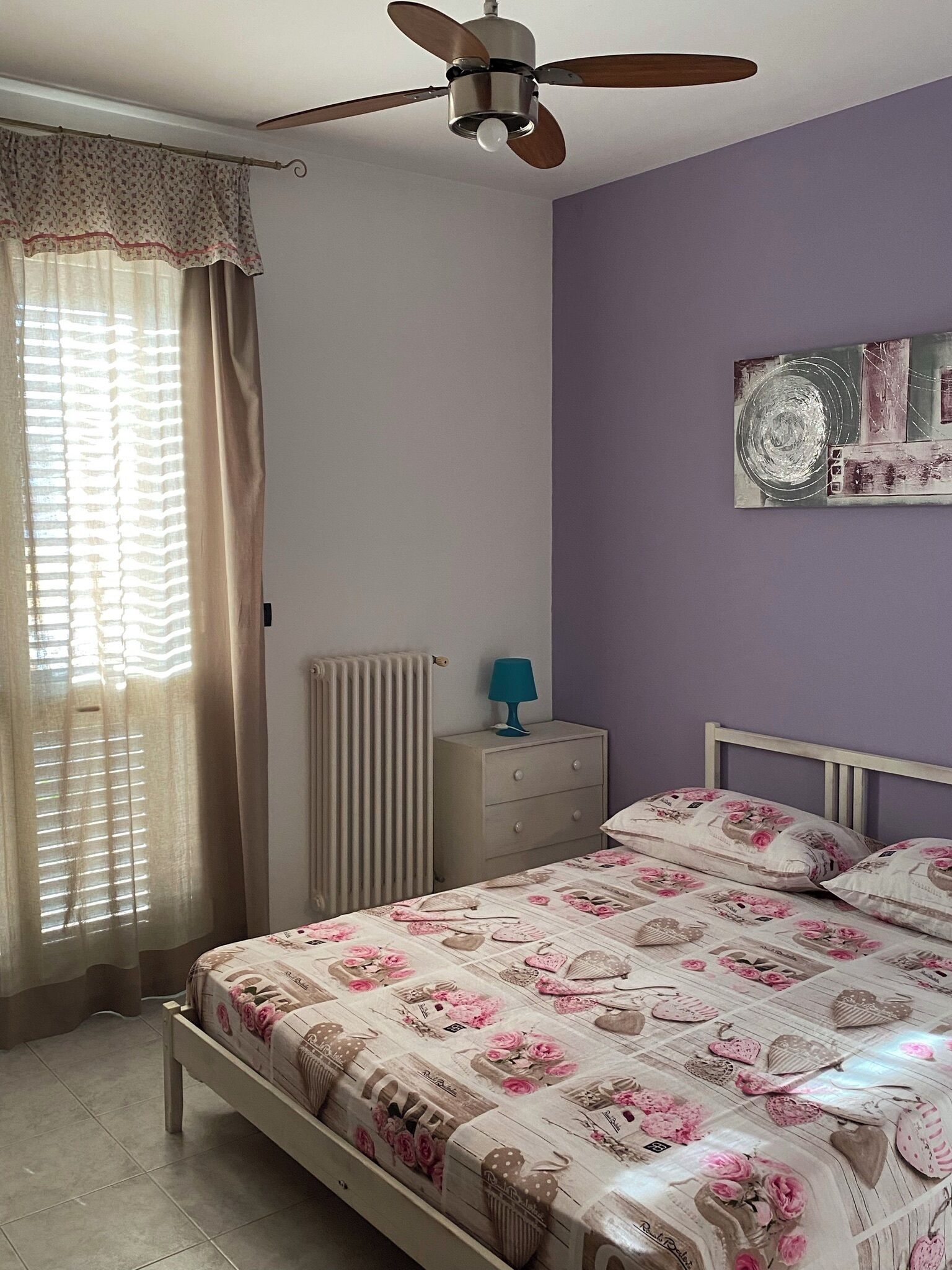 Une chambre à coucher comprenant un lit, un ventilateur de plafond, un radiateur, une table de chevet et un mur orné d’un tableau.