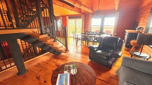 A wooden interior with a staircase, a black leather sofa, a round coffee table with a book, and a dining area with chairs and a table.