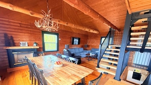 A cozy cabin interior with a wooden dining table, chairs, a fireplace, a sofa, and a staircase.