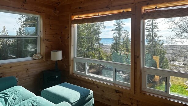 A cabin with wooden walls, a bed, a bench, and large windows offering a view of trees and a body of water.
