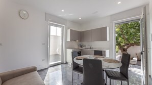 Une cuisine moderne dotée d’appareils électroménagers intégrés, un coin repas avec une table ronde et des chaises, et une grande porte-fenêtre coulissante donnant sur un balcon agrémenté d’un arbre.