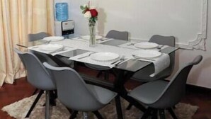 Un coin repas avec une table dressée pour quatre personnes, des chaises grises, un lustre, un distributeur d’eau et un tableau encadré au mur.