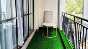 Un balcon étroit doté d’un sol recouvert d’un gazon artificiel vert, d’une chaise blanche et offrant une vue sur les arbres à travers la fenêtre.