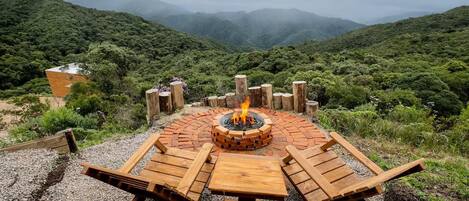 A circular wooden fire pit with a fire, surrounded by wooden benches and a table, set on a gravel surface with a mountainous backdrop.