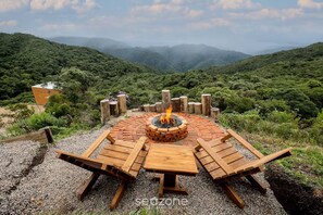 A circular wooden fire pit with a fire, surrounded by wooden benches and a table, set on a gravel surface with a mountainous backdrop.
