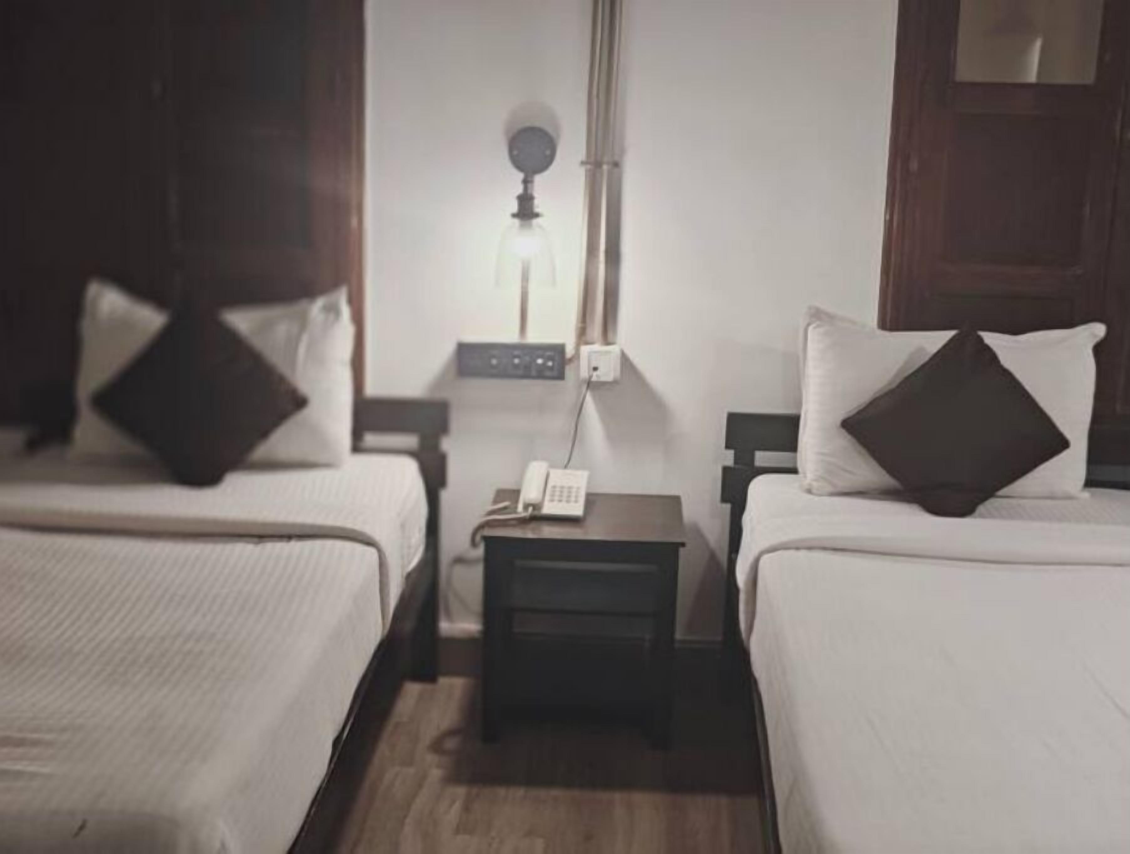 Habitación de hotel con dos camas individuales, cada una con una almohada y una mesita de noche. Hay una mesita de noche entre las camas con una lámpara y un teléfono.