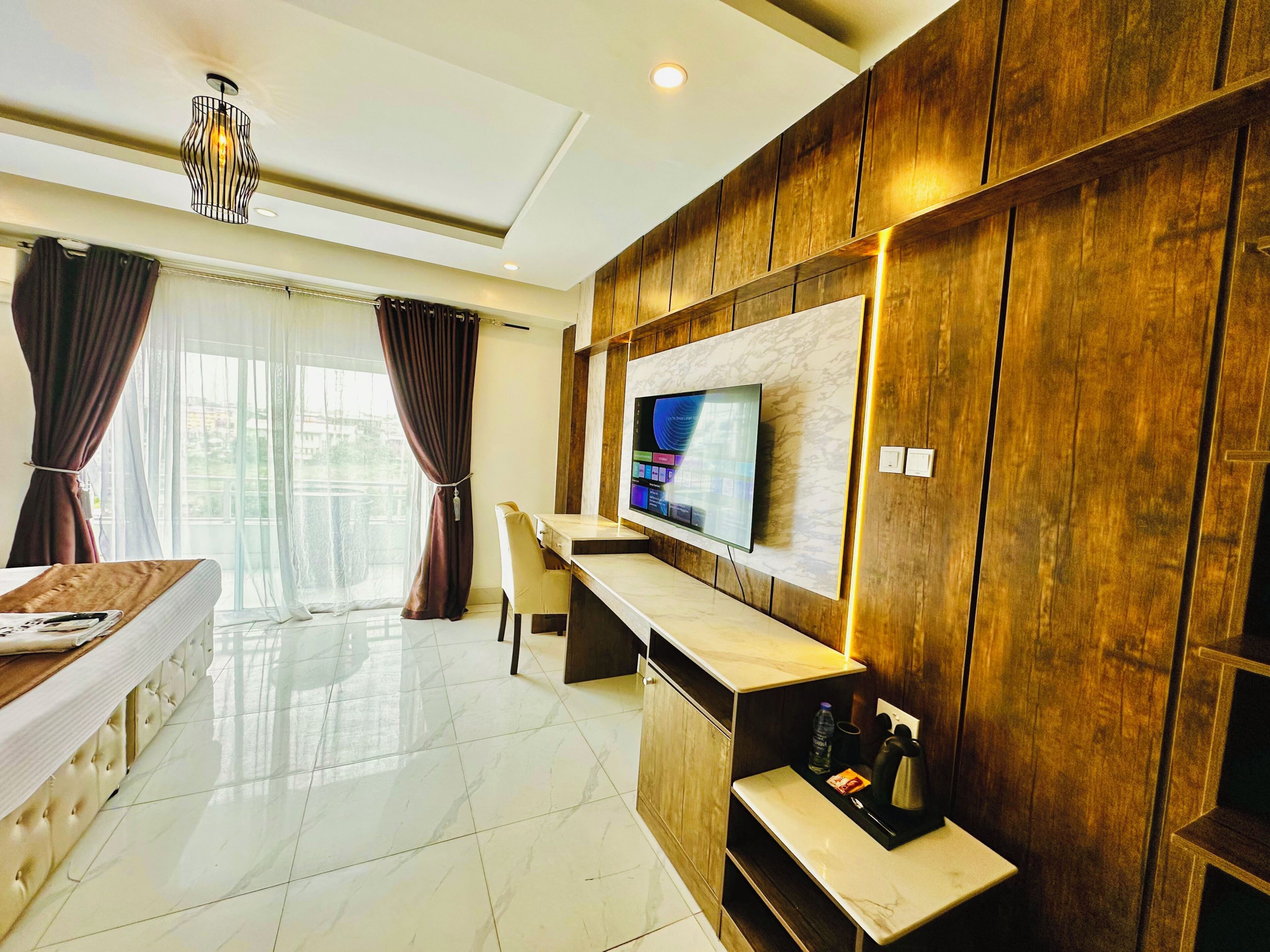 Habitación de hotel moderna con una televisión de pantalla plana montada en una pared de paneles de madera, una cama cómoda con cabecera, un escritorio pequeño con una silla y un ventanal con cortinas.