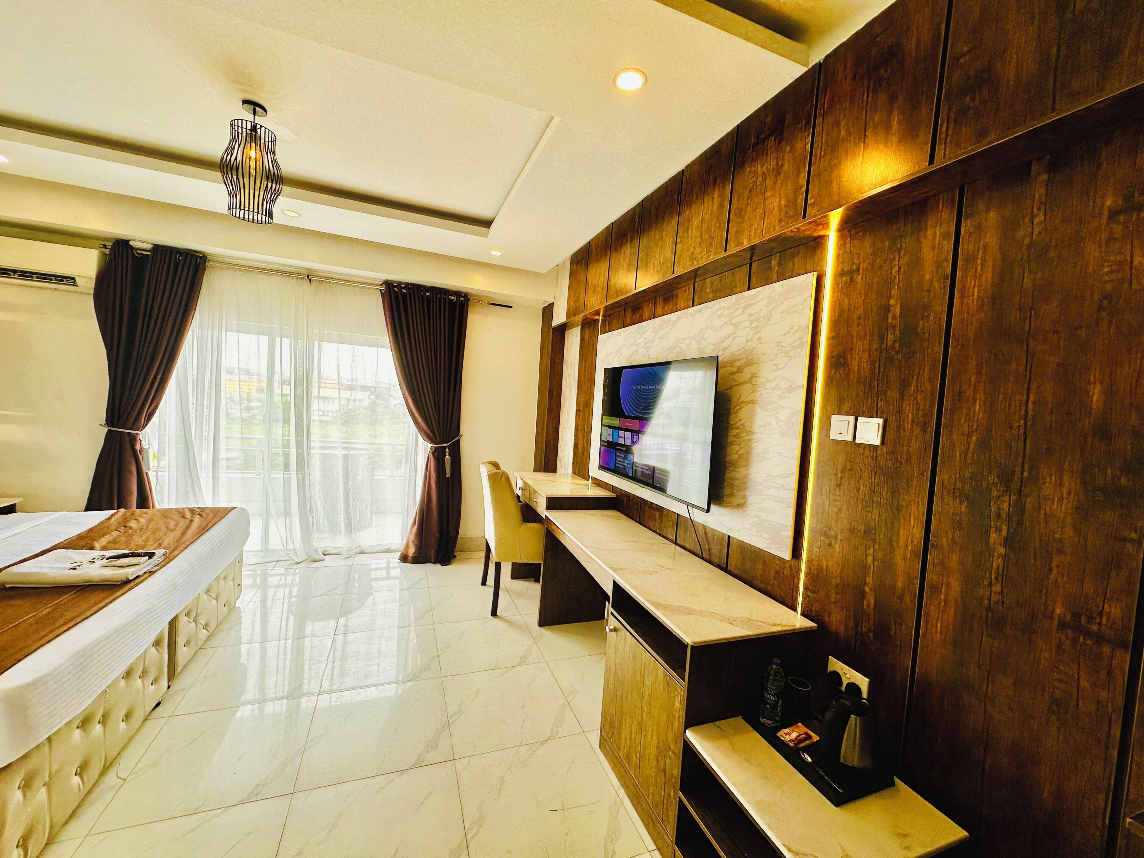 Una habitación de hotel moderna con un televisor grande montado en una pared revestida de paneles de madera, una zona de comedor con mesa y sillas, y un ventanal con cortinas.