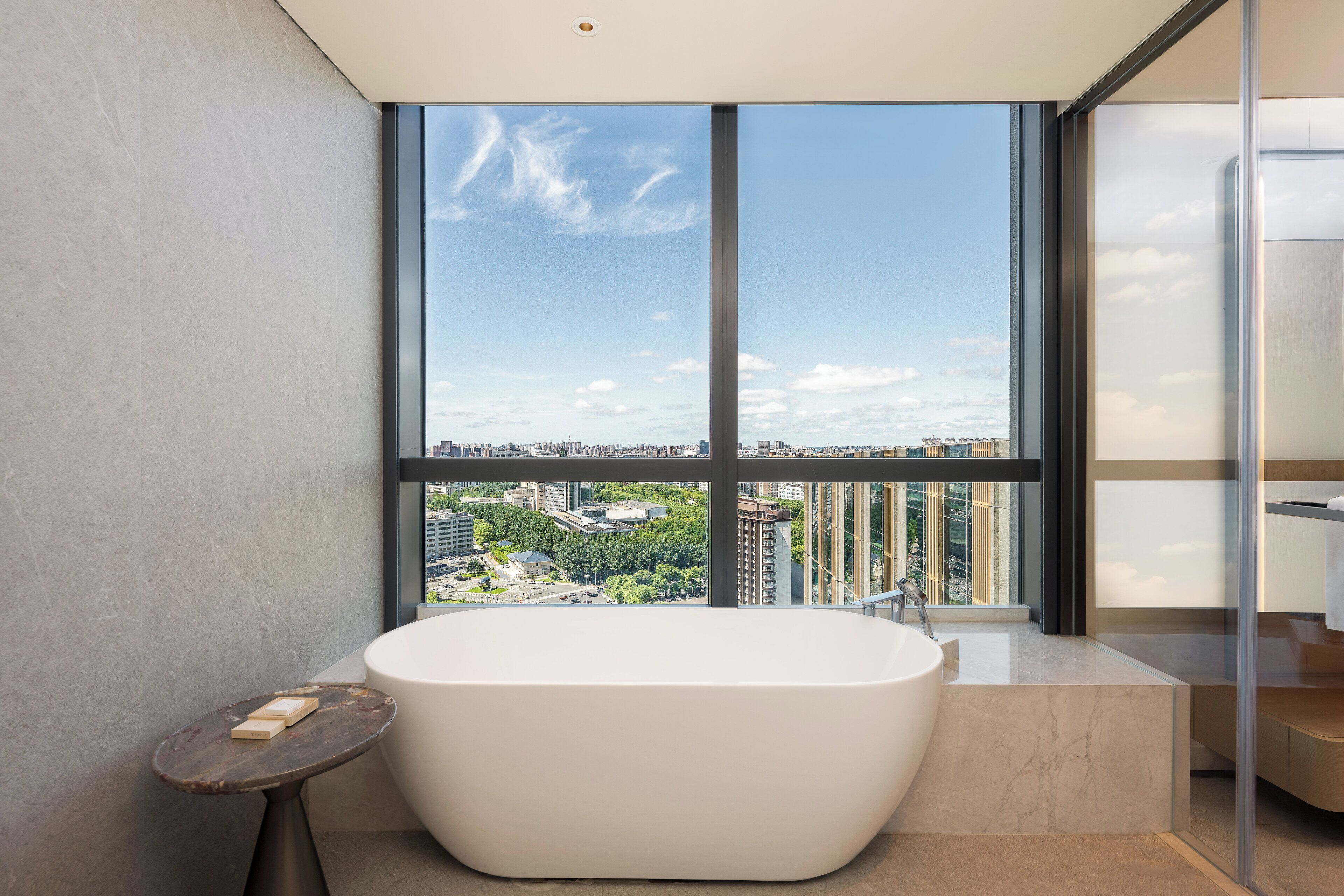 A modern bathroom with a freestanding bathtub, a small table with toiletries, and a large window offering a city view.