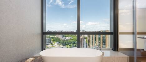 A modern bathroom with a freestanding bathtub, a small table with toiletries, and a large window offering a city view.