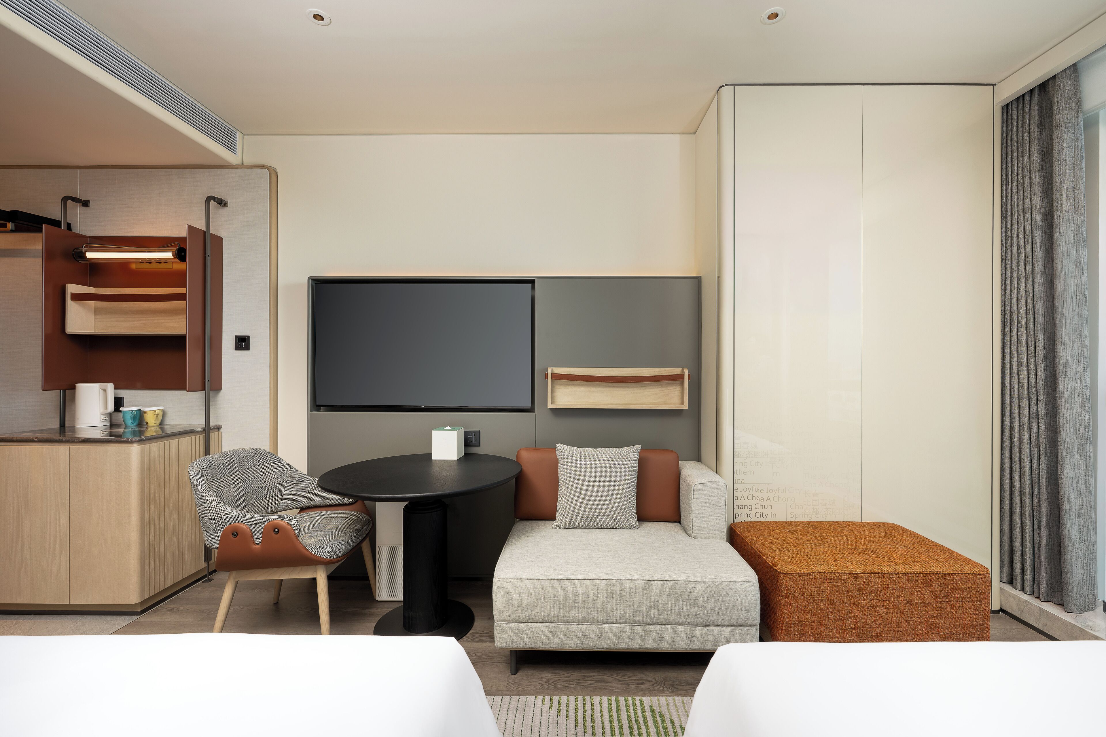 A modern hotel room with a sofa, a small table, a chair, a TV, and a large window.