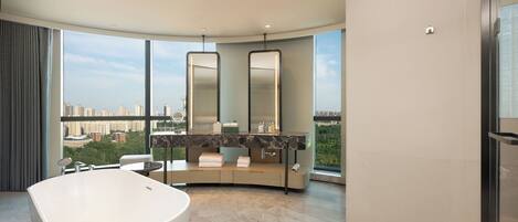 Un baño moderno con una bañera independiente, un tocador curvo con un espejo grande y vistas de la ciudad a través de amplias ventanas.
