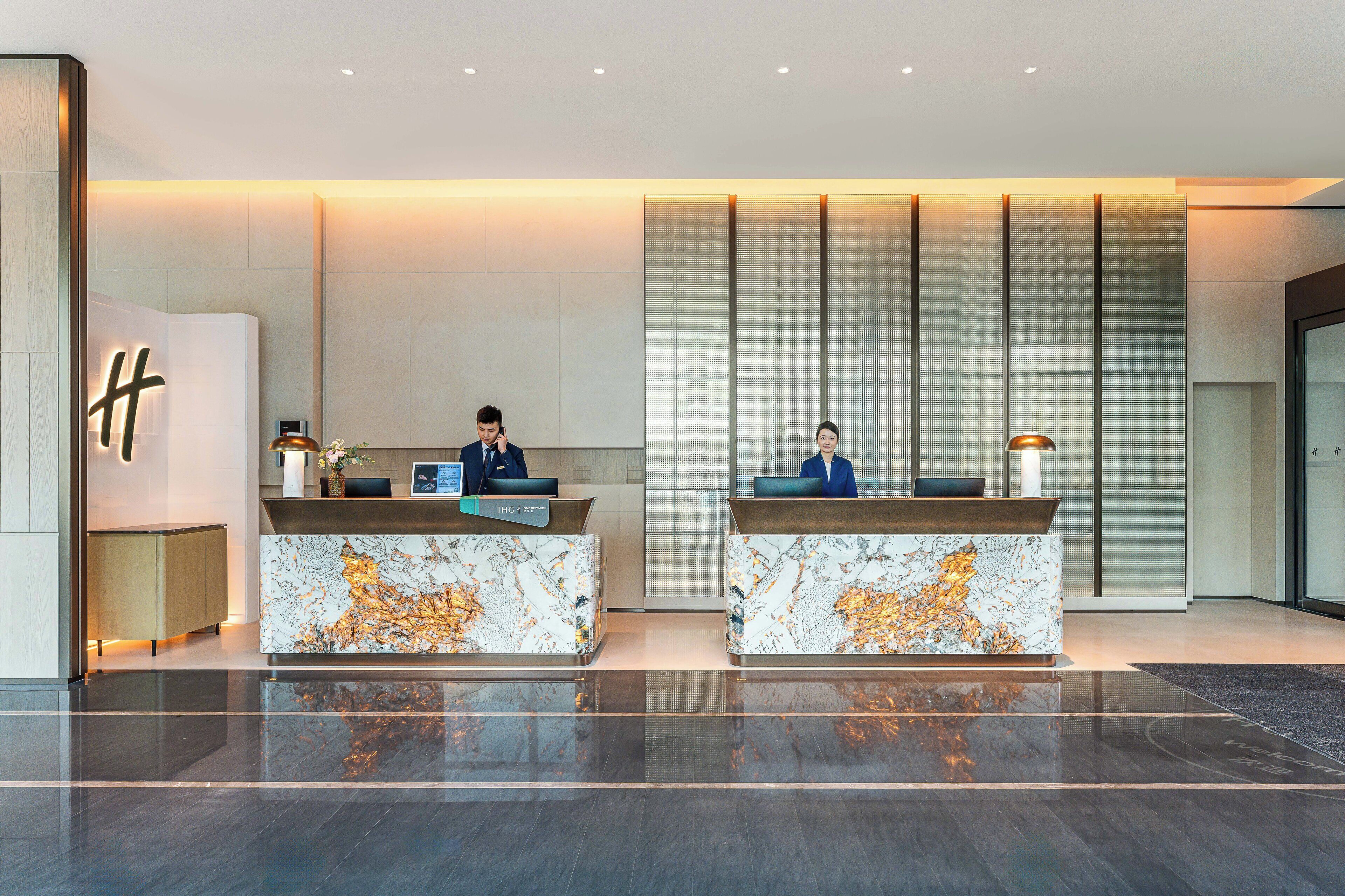 A modern hotel reception area with marble counters, two receptionists, and a large illuminated 'H' sign.