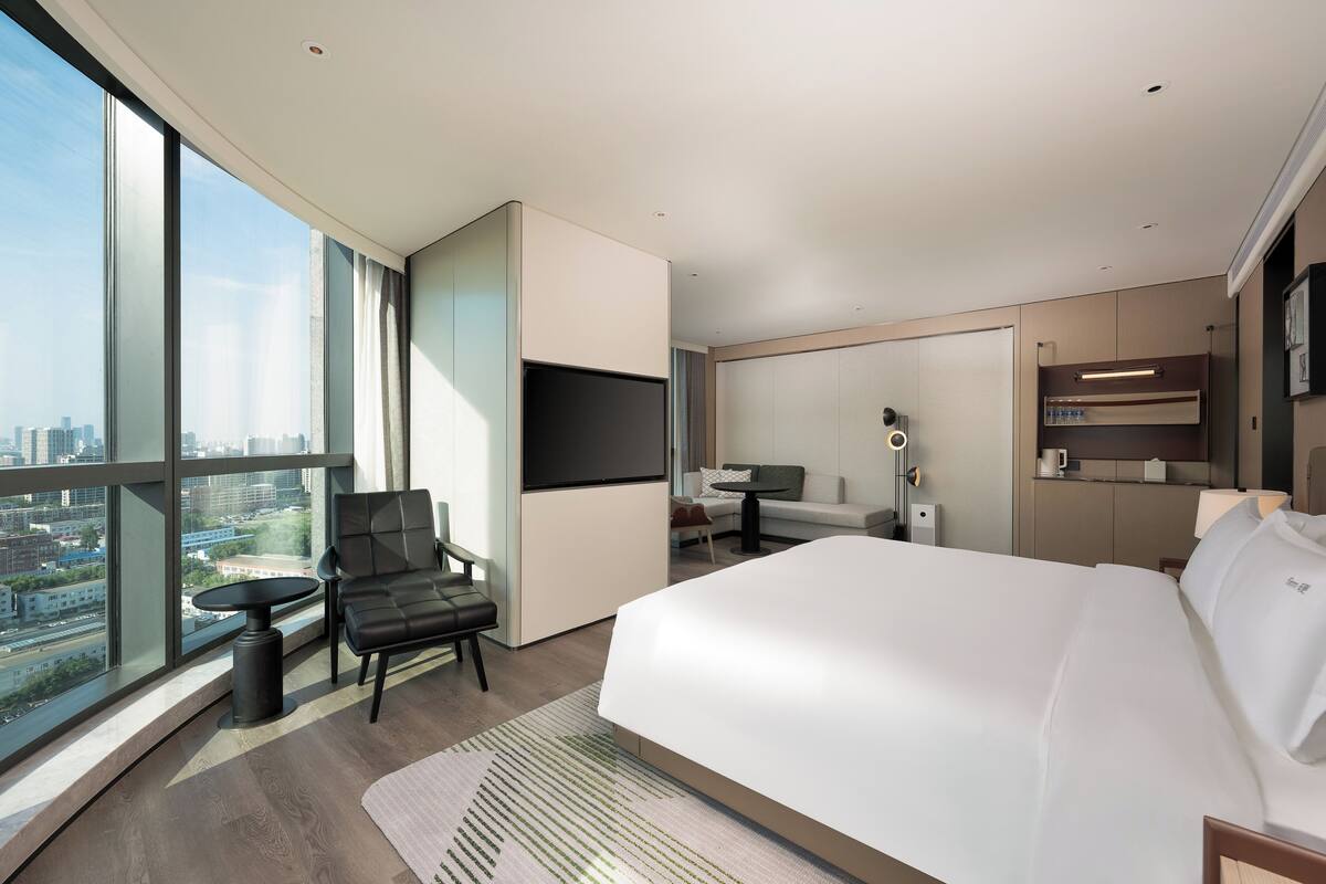 Une chambre d’hôtel moderne dotée d’un grand lit, d’un coin salon avec un fauteuil et une table, d’un téléviseur à écran plat, d’un bureau avec une lampe et offrant une vue sur la ville grâce à de grandes fenêtres.