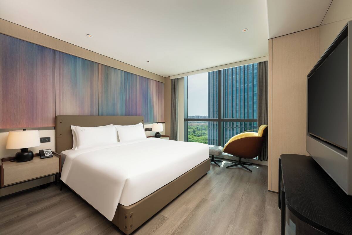 Une chambre d’hôtel moderne dotée d’un grand lit, d’une télévision à écran plat, d’un fauteuil et offrant une vue sur la ville.