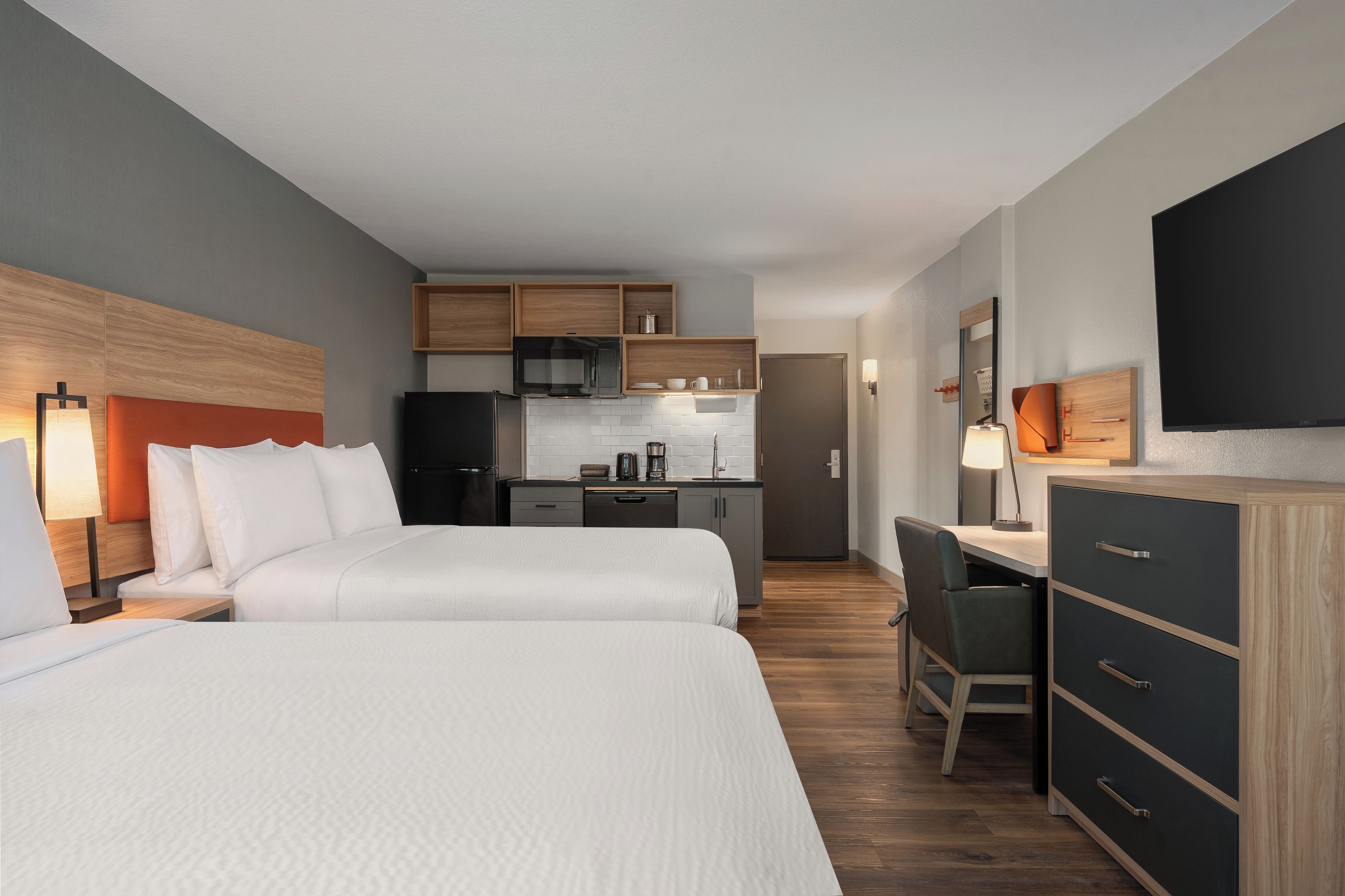 A hotel room with two beds, a kitchenette, a desk, and a TV.