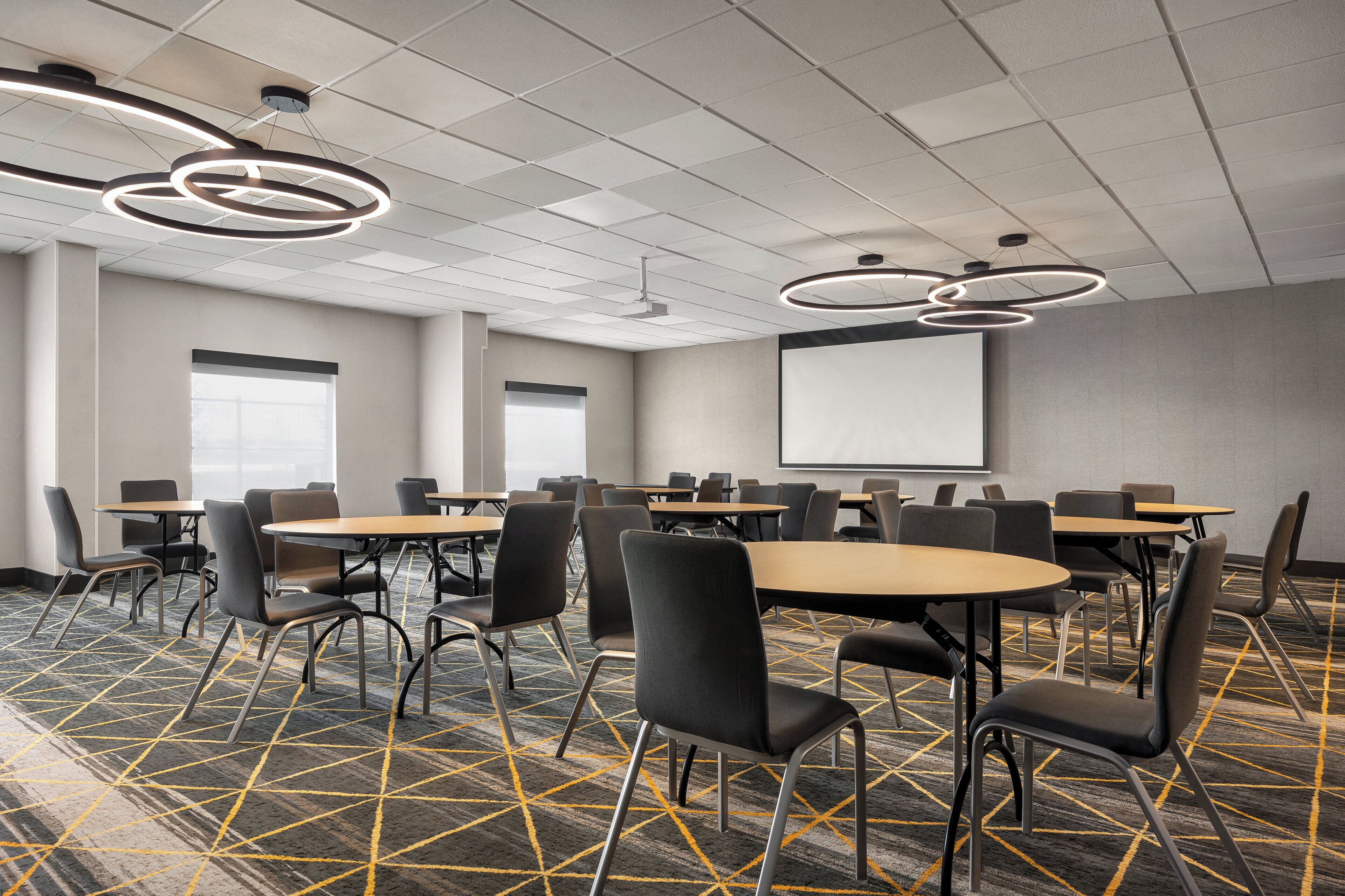 Une salle de conférence équipée de plusieurs tables et chaises, d’un écran de projection et d’un tapis à motifs.