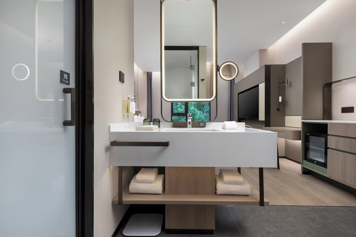 A modern bathroom with a large mirror, a sink, and a toilet. There is a television and a cabinet in the background.