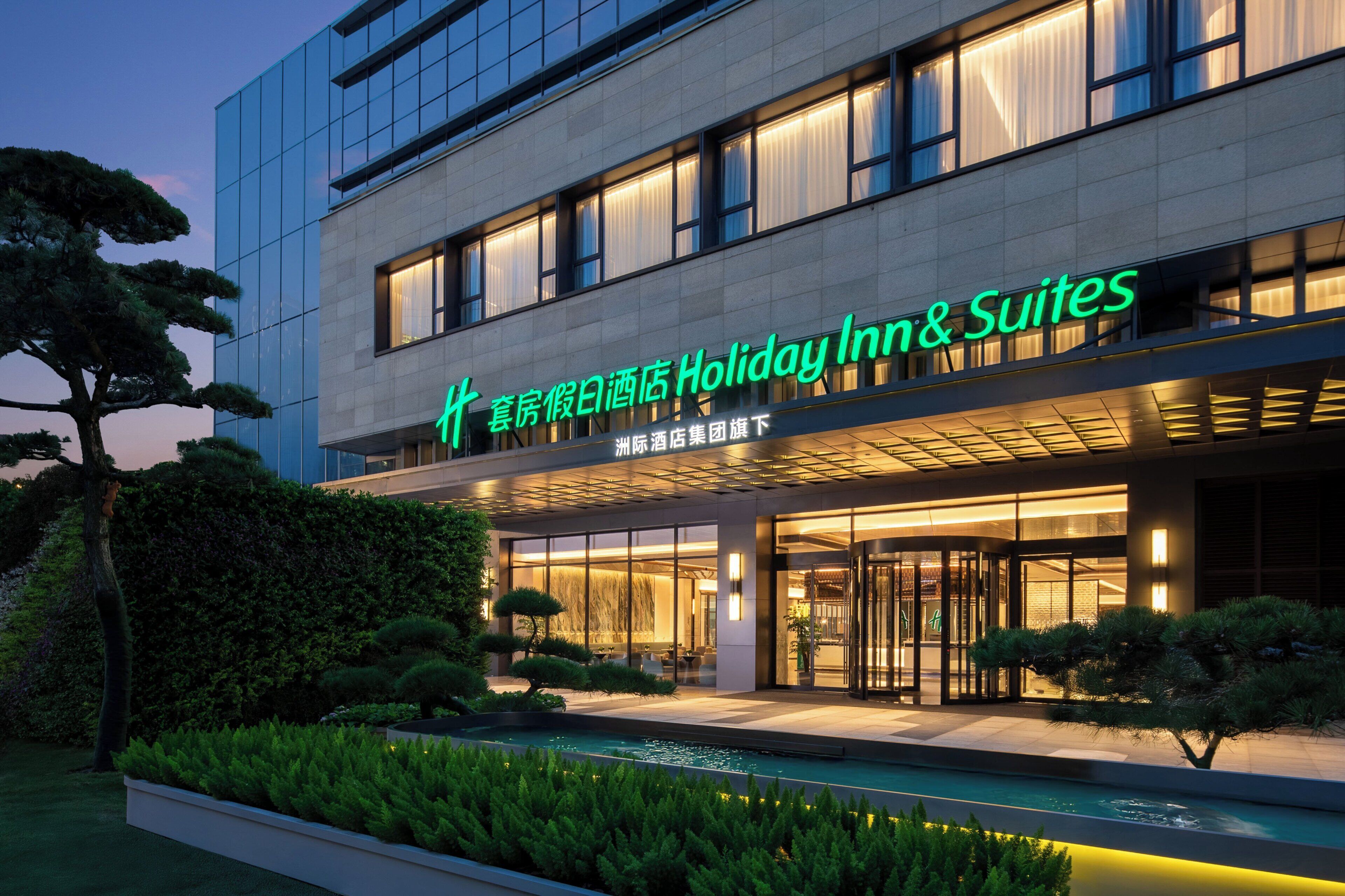 A modern hotel with a glass facade, illuminated signage, and a landscaped entrance area.