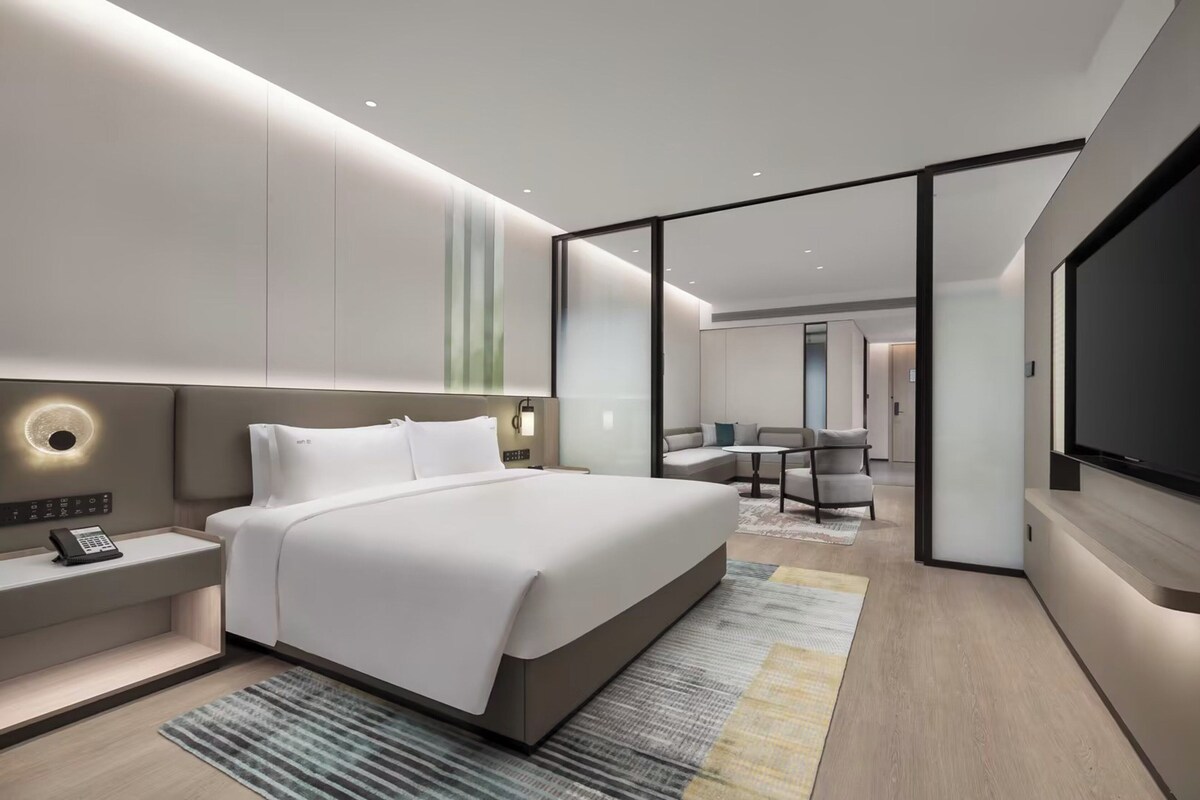 A modern hotel room with a large bed, bedside controls, a flat-screen TV, and a seating area with a table and chairs.