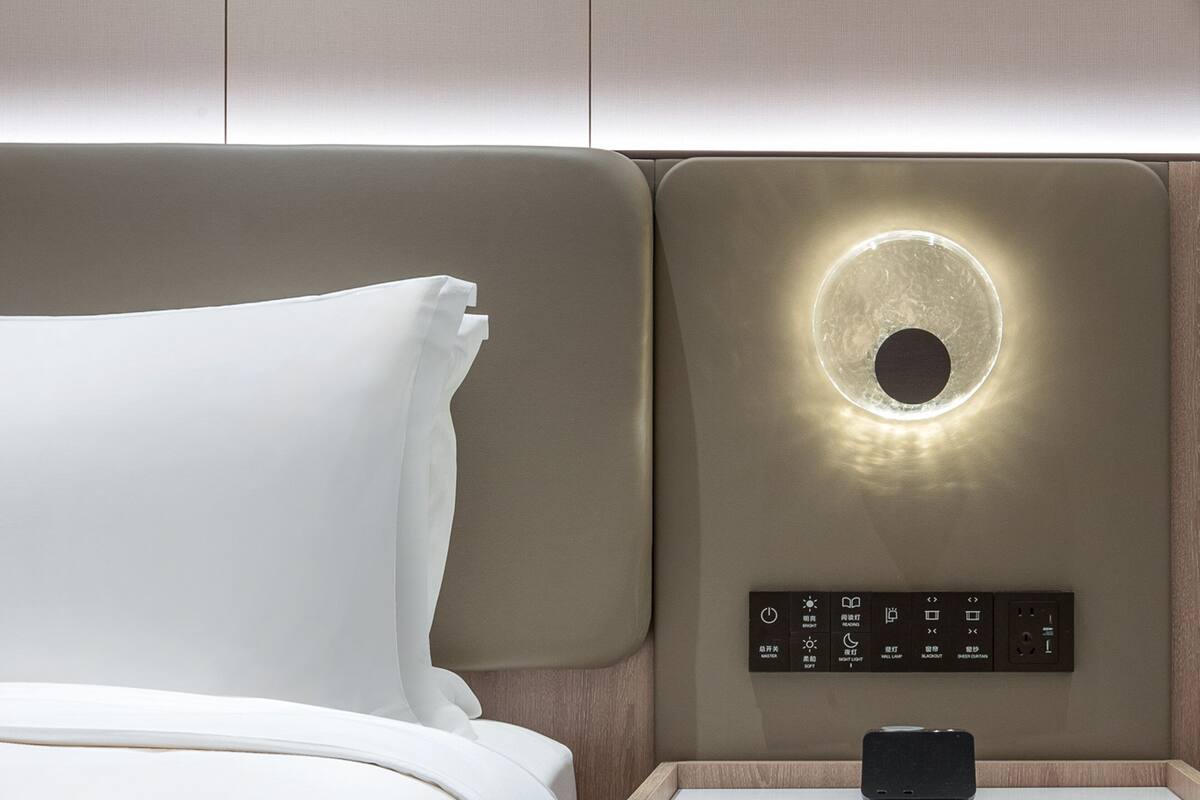 A modern hotel room with a bed, a control panel with various buttons, and a wall-mounted light fixture.