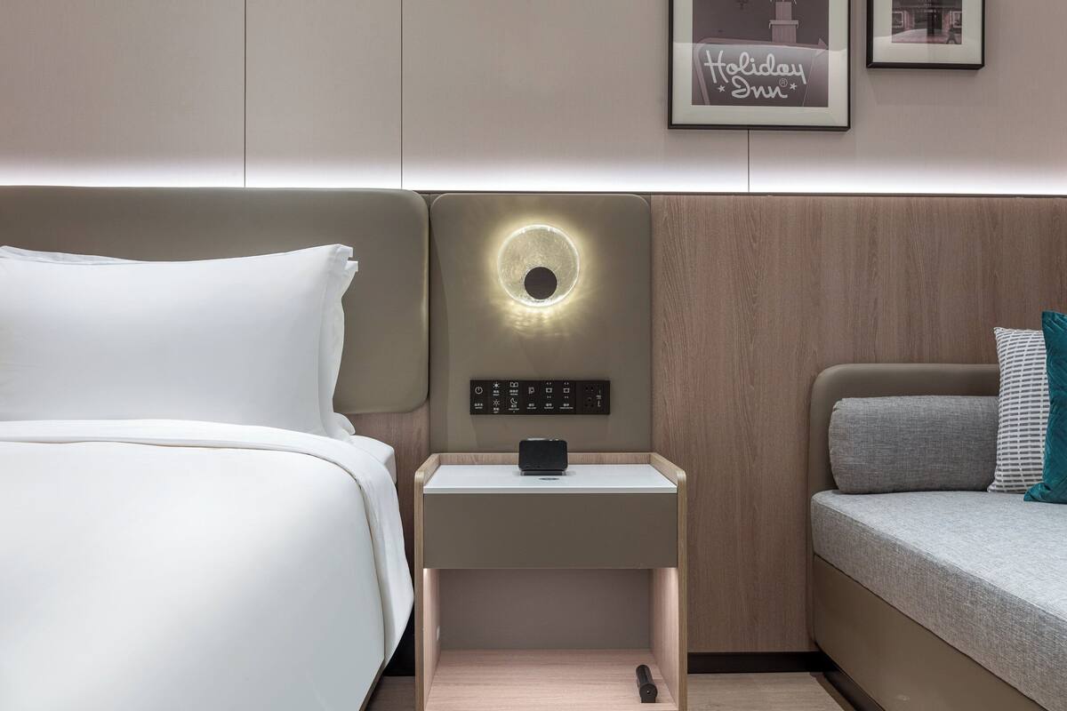 A modern hotel room with a bed, a bedside table with a built-in lamp, a control panel, and a framed picture on the wall.