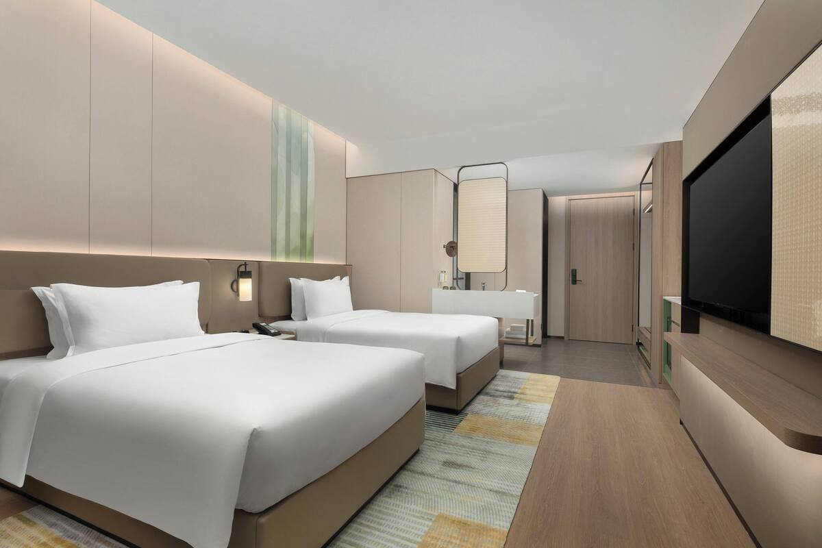 A modern hotel room with two beds, a large flat-screen TV, a bathroom with a mirror, and a wooden floor.