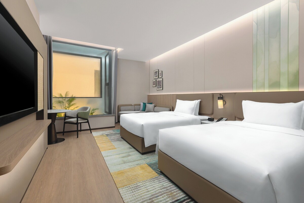 A modern hotel room with two beds, a desk, and a large window.