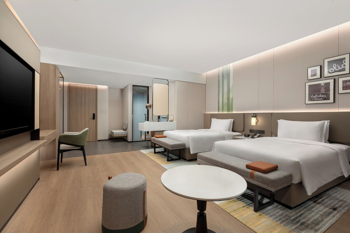 A modern hotel room with a large bed, a smaller bed, a desk, a chair, a round table, and a TV.