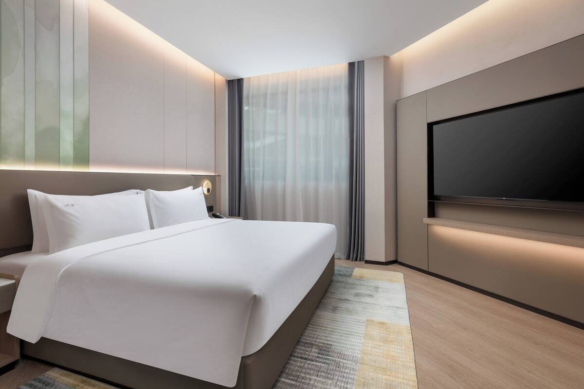A modern hotel room with a large bed, a flat-screen TV, and a large window with curtains.