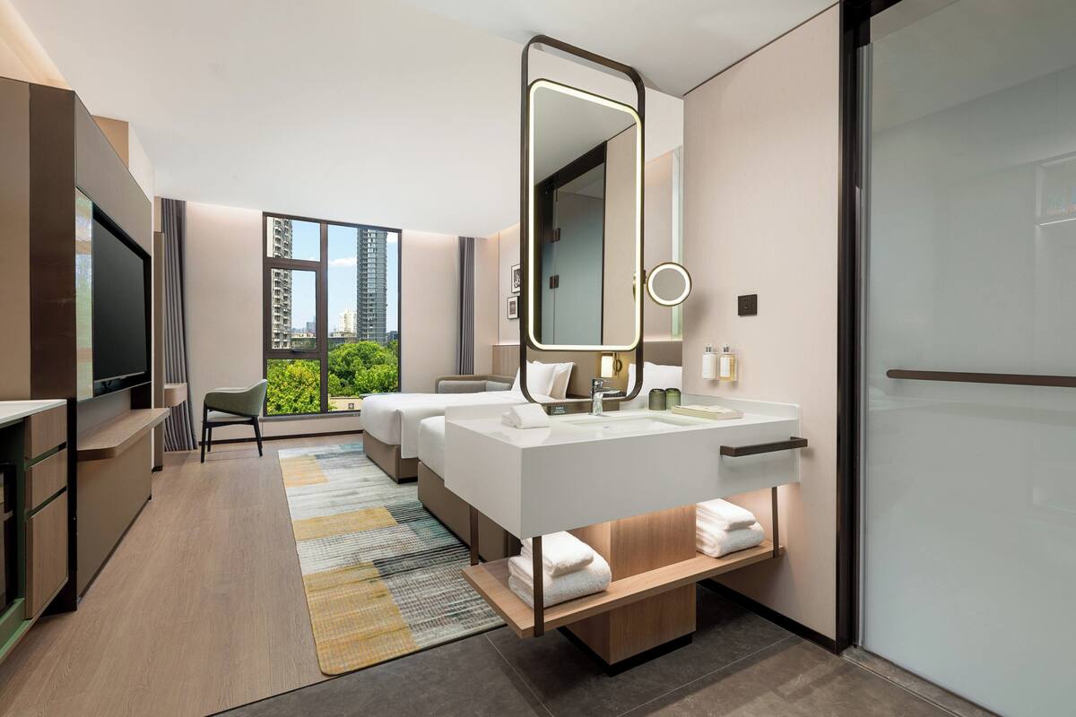 A modern hotel room with a large bed, a separate sitting area, a TV, and a bathroom with a large mirror and sink.