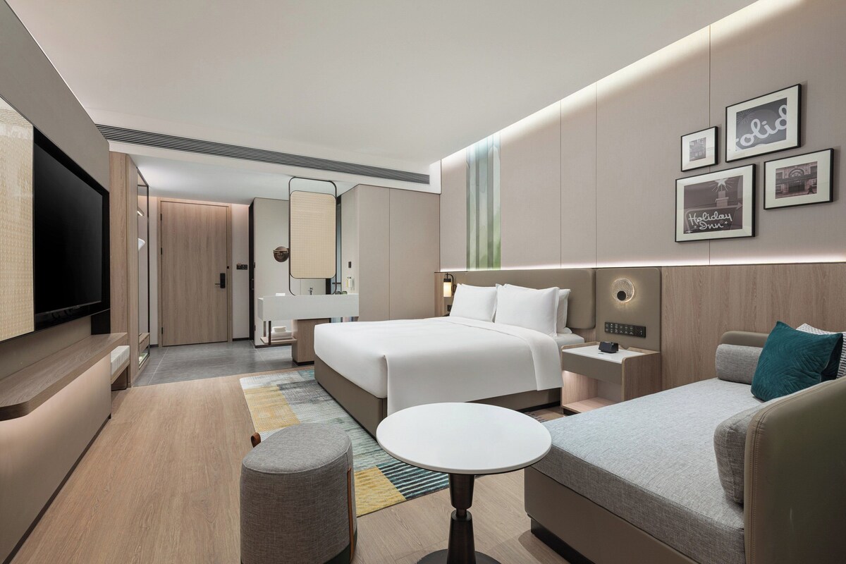 A modern hotel room with a large bed, a sofa, a small table, and a TV. There are framed pictures on the wall and a mirror above the bed.