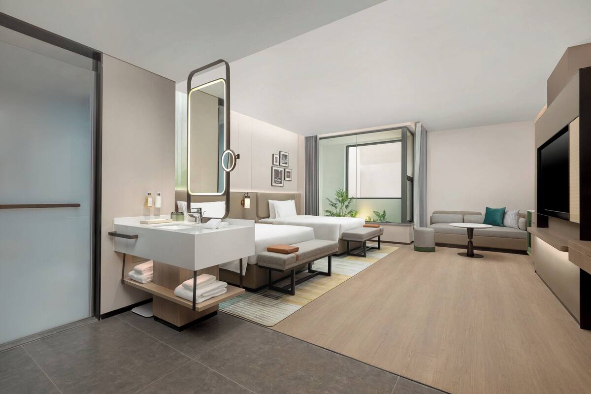 A modern hotel room with a large bed, a sofa, a TV, and a bathroom with a mirror and sink.