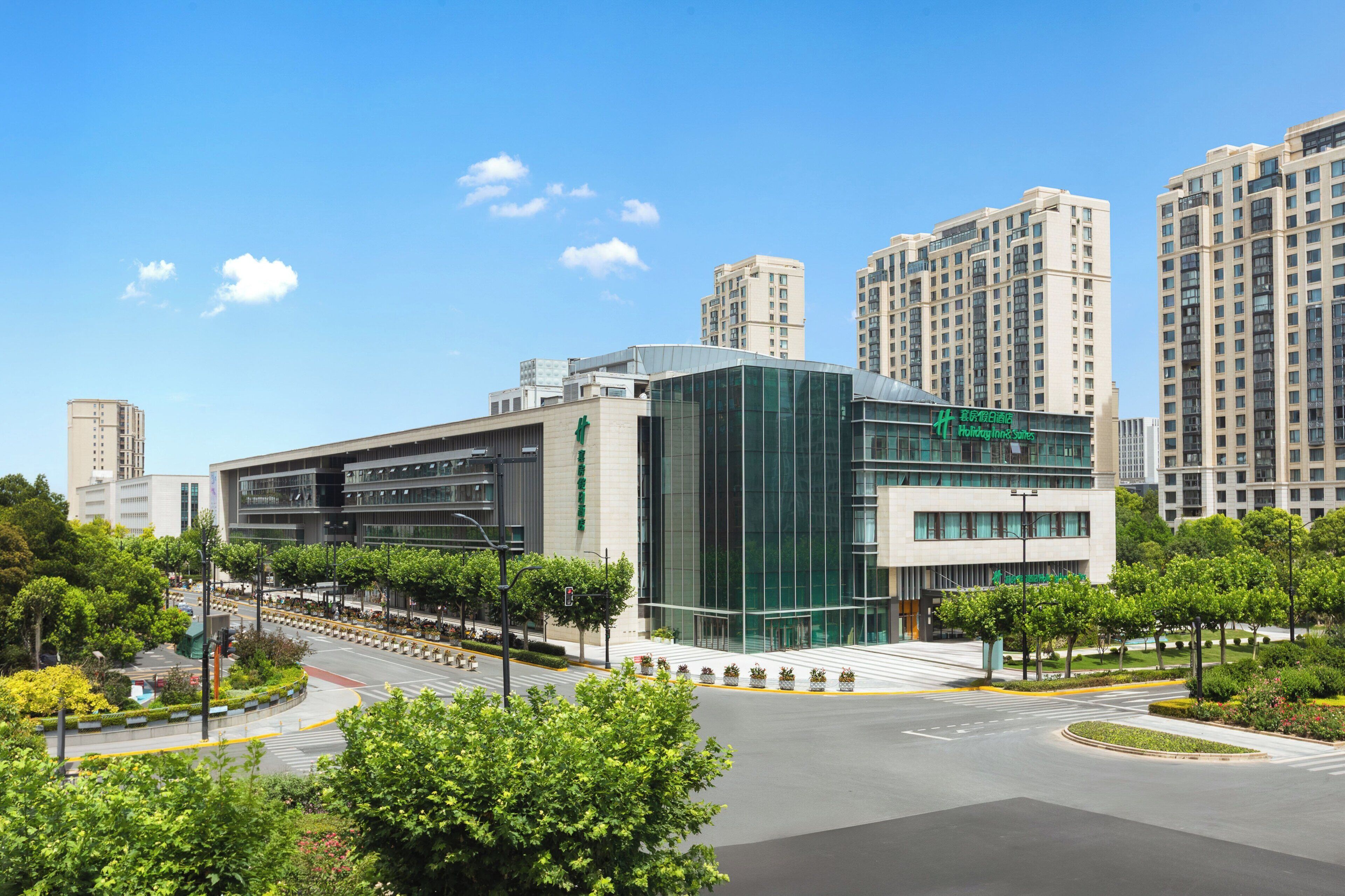 A modern hotel with a glass facade, surrounded by greenery and high-rise buildings.