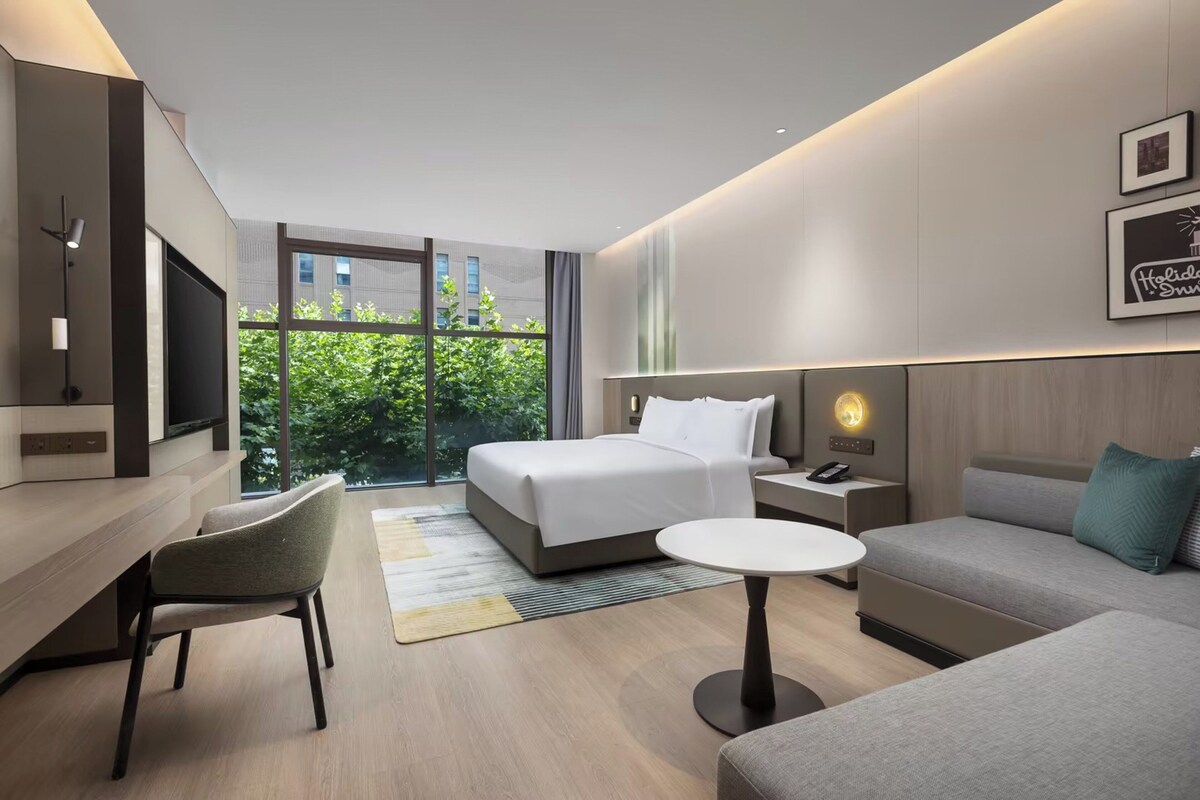 A modern hotel room with a bed, a sofa, a desk, and a TV. There is a window with a view of greenery outside.