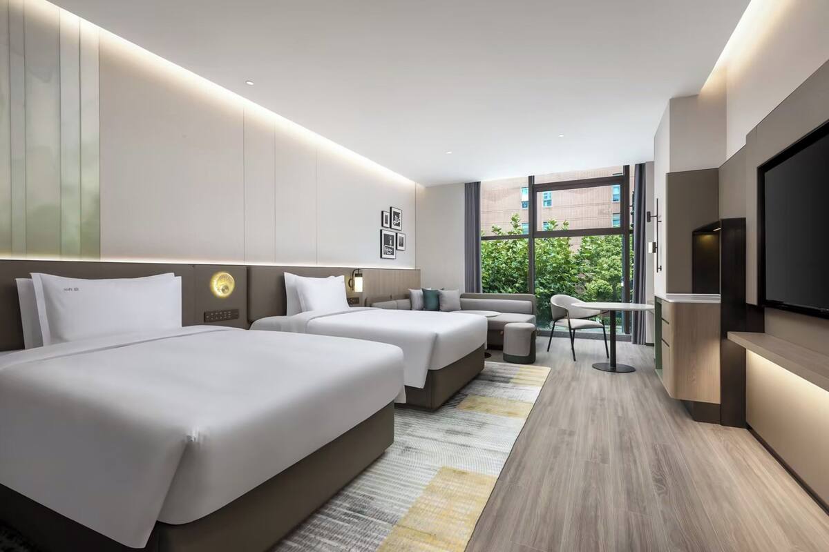 A modern hotel room with two beds, a sofa, a TV, and a view of greenery outside.
