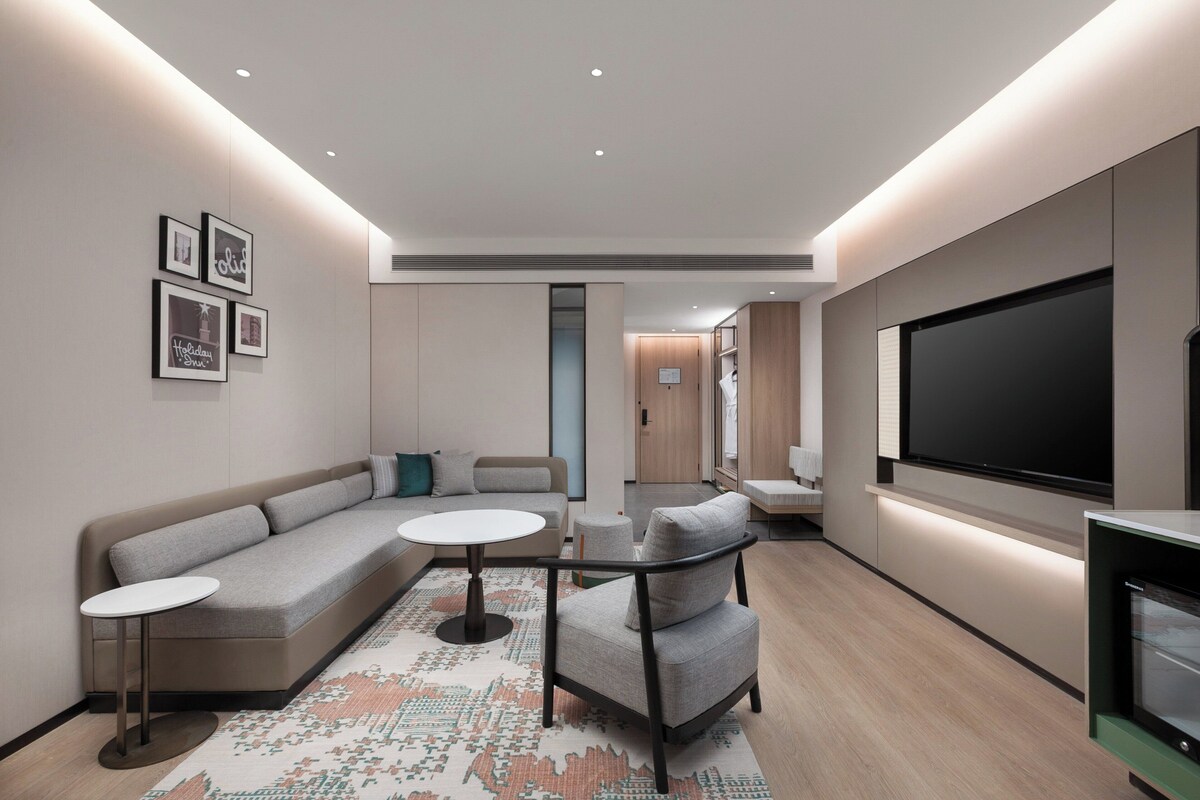 A modern living room with a sectional sofa, a small round table, a chair, a flat-screen TV, and a large rug.