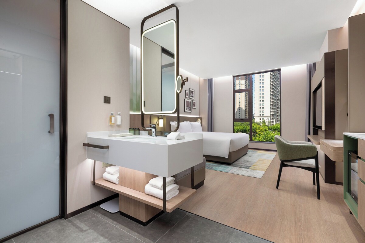 A modern hotel room with a large bed, a separate bathroom with a vanity, a chair, and a view of the city through large windows.