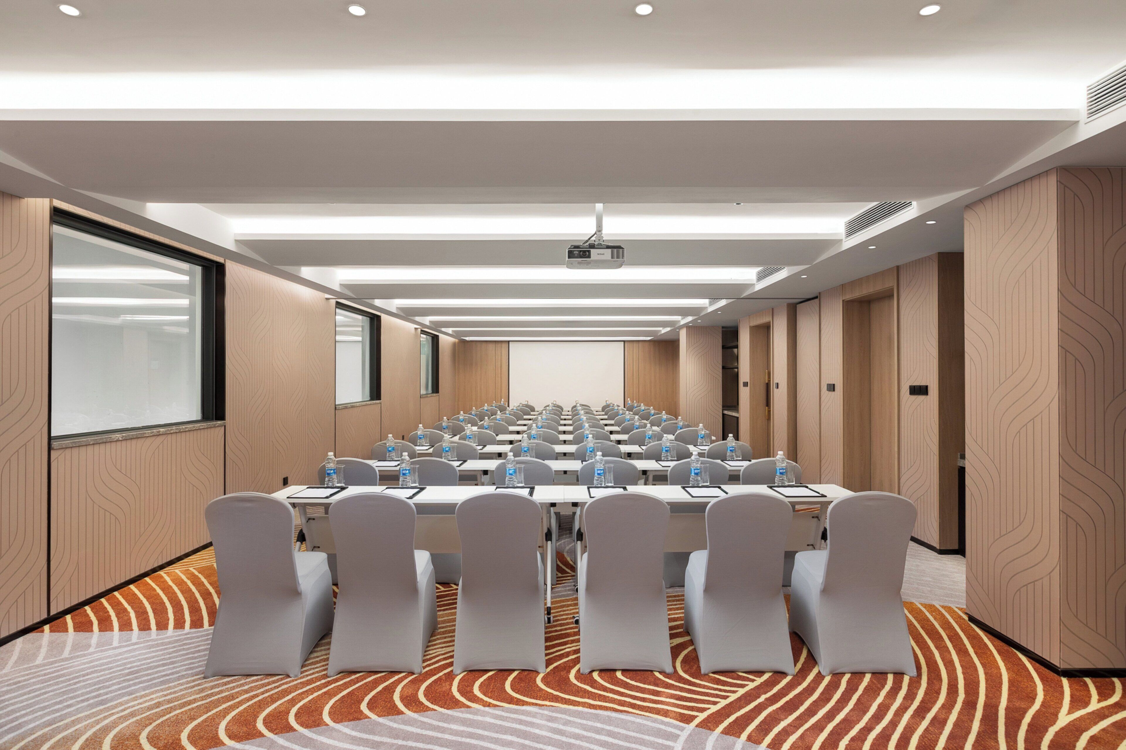 Een congreszaal met rijen tafels en stoelen, een beamer en een groot scherm.