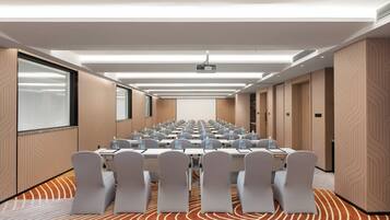 Een congreszaal met rijen tafels en stoelen, een beamer en een groot scherm.