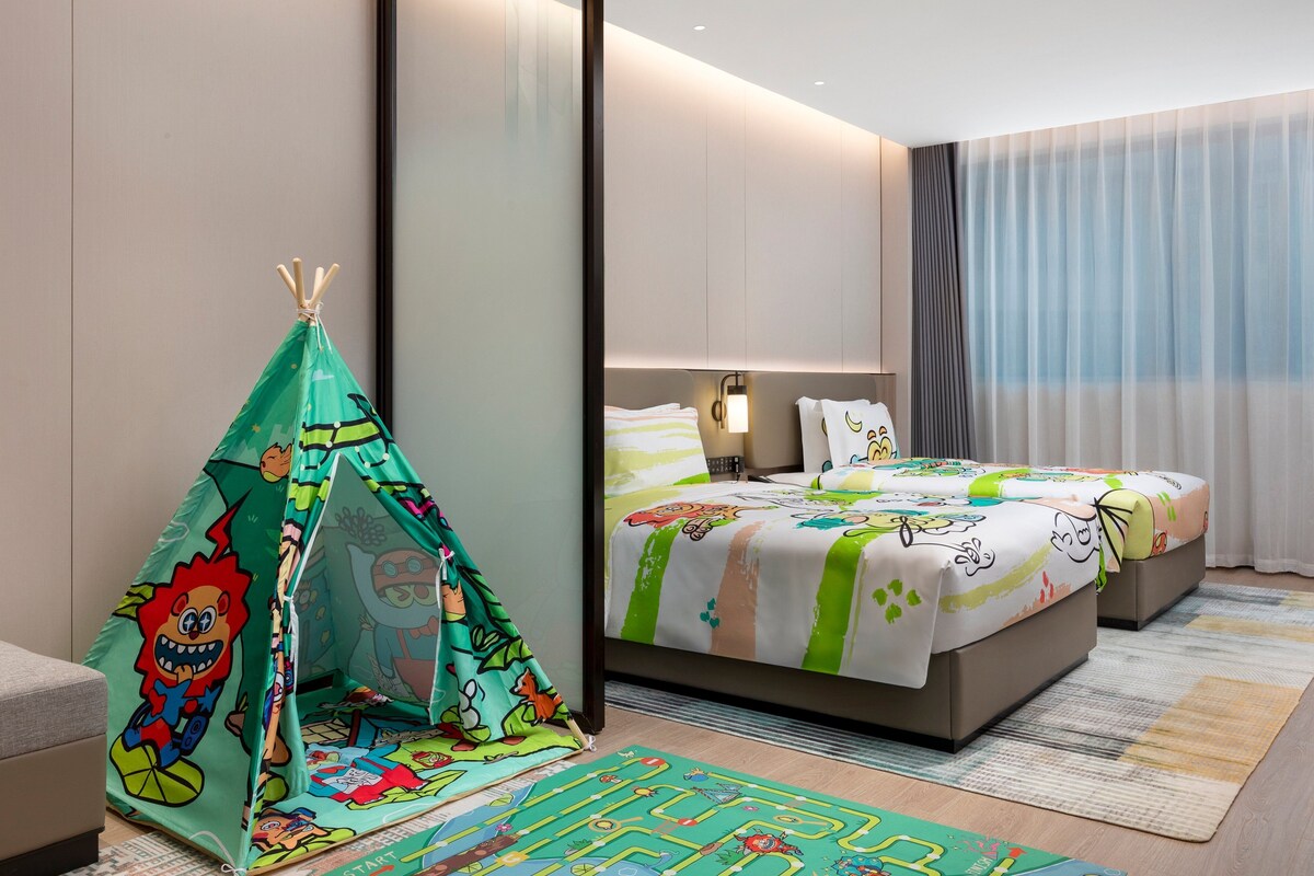 A hotel room with a bed, a green tent with cartoon characters, and a large window with curtains.