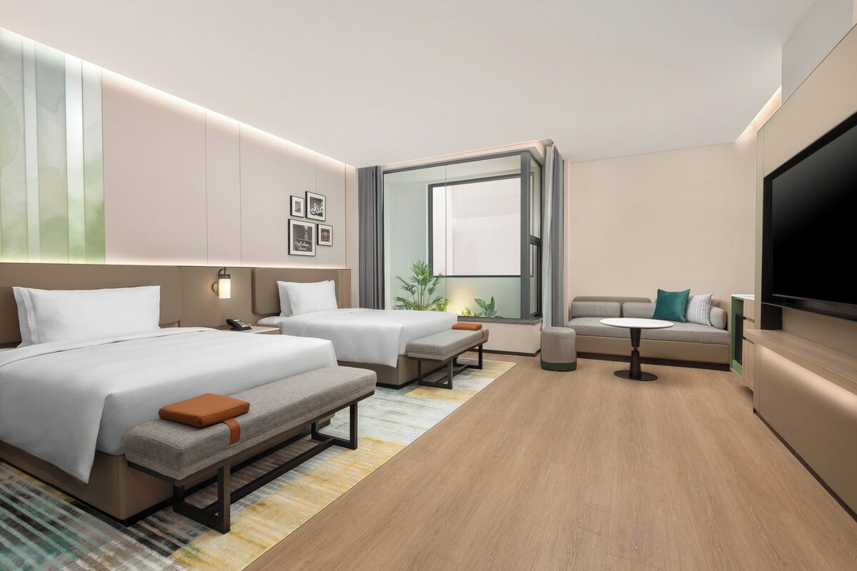 A modern hotel room with two beds, a sofa, a TV, and a large window.