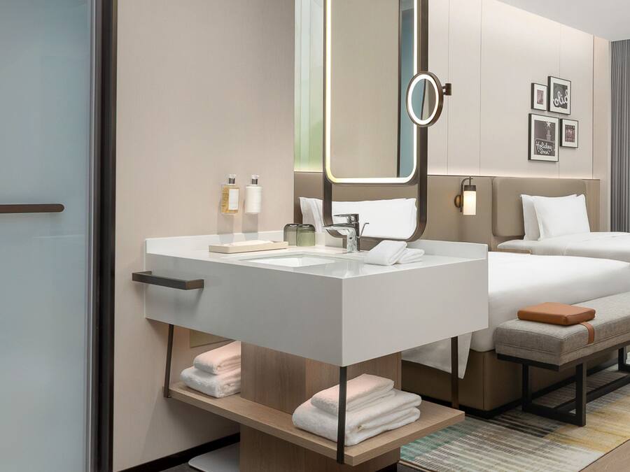 A modern hotel room with a large bed, a bedside table, a vanity with a mirror, and a small seating area.