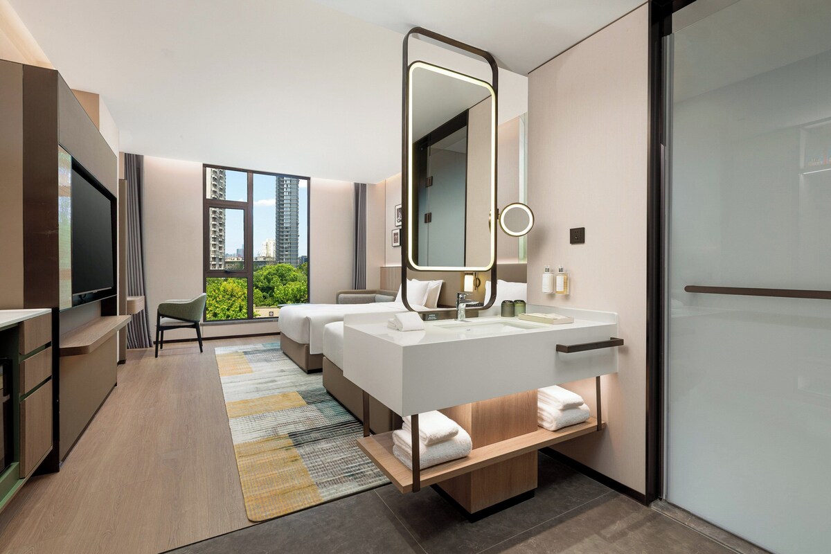 A modern hotel room with a large bed, a separate sitting area, a TV, and a bathroom with a large mirror and sink.