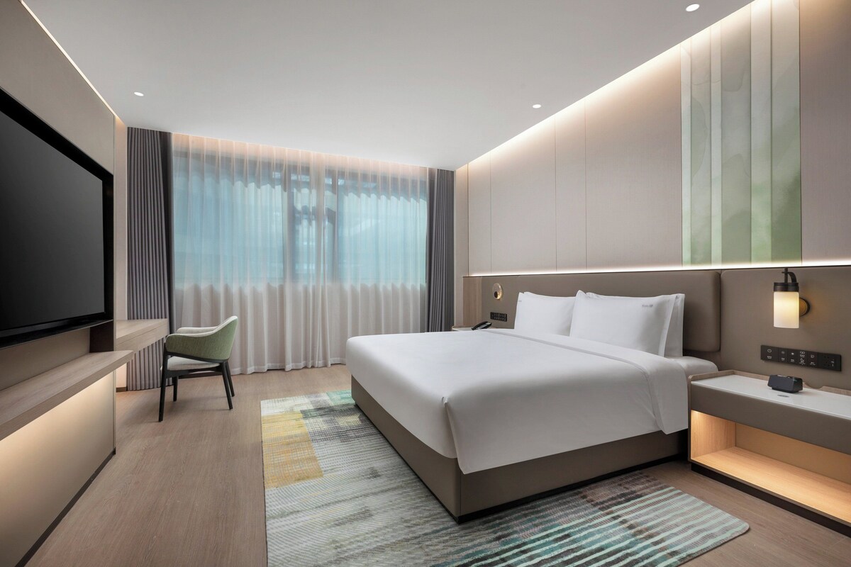 A modern hotel room with a large bed, bedside tables, a flat-screen TV, and a view of the city through large windows.